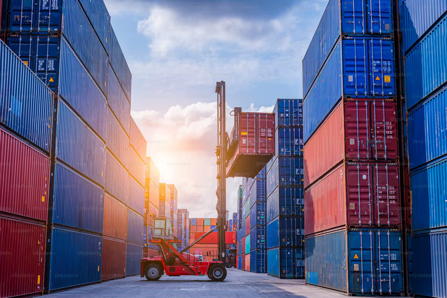 “A reach stacker lifting a red shipping container between tall stacks of blue and red containers at a terminal”