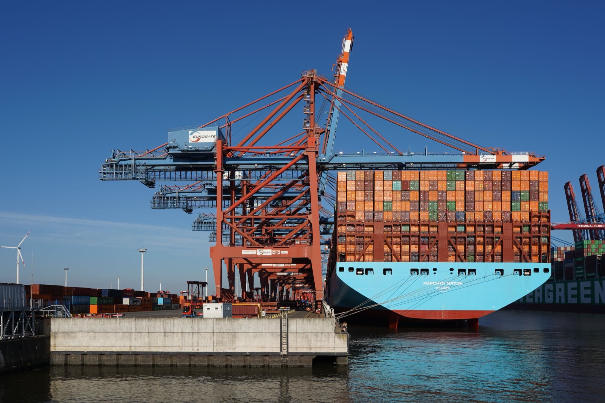 A container ship loaded with containers, berthed under ship-to-shore cranes at a container terminal
