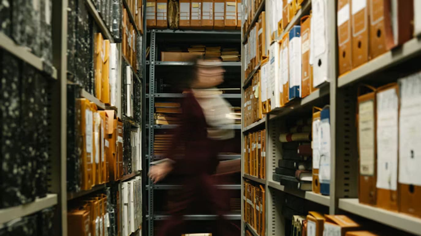 A narrow archive aisle lined with shelves of orange and grey file boxes, a person in a dark red suit blurred in motion as they walk past.