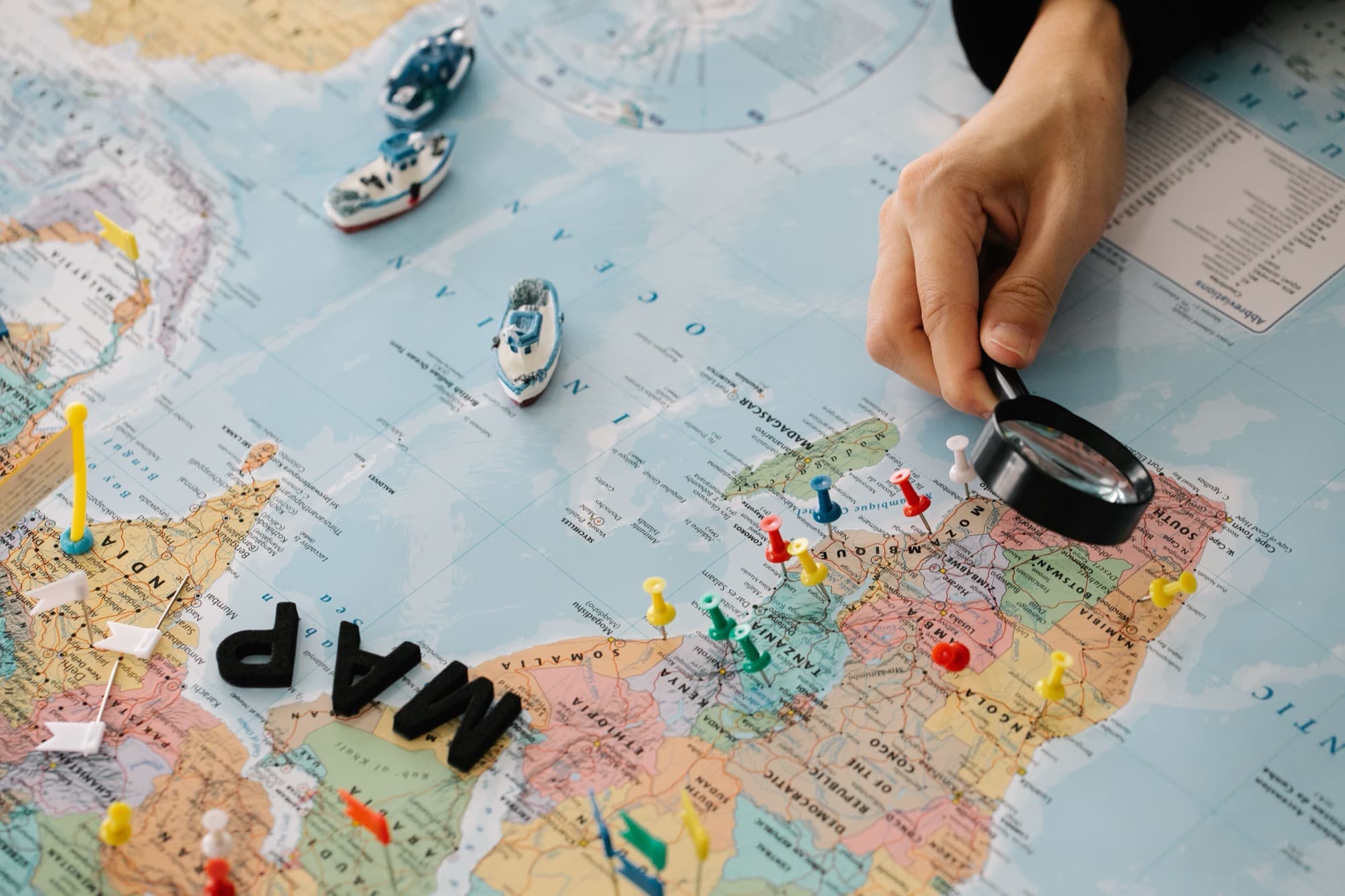 A hand holding a magnifying glass over a world map, with toy cargo ships on the Indian Ocean and coloured pins marking places across East and Southern Africa