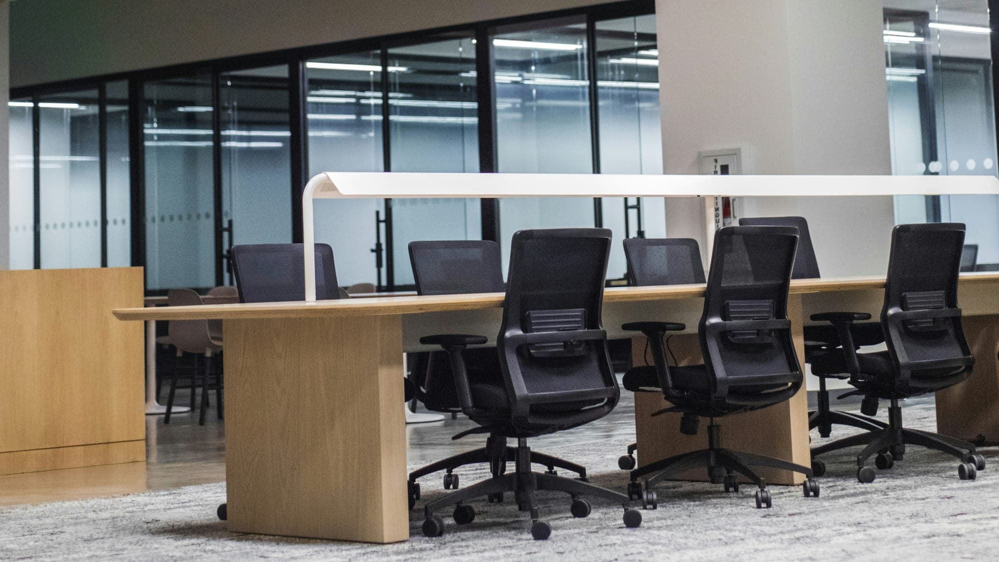 An empty open-plan office: black mesh chairs at a long oak desk, glass-walled meeting rooms behind.