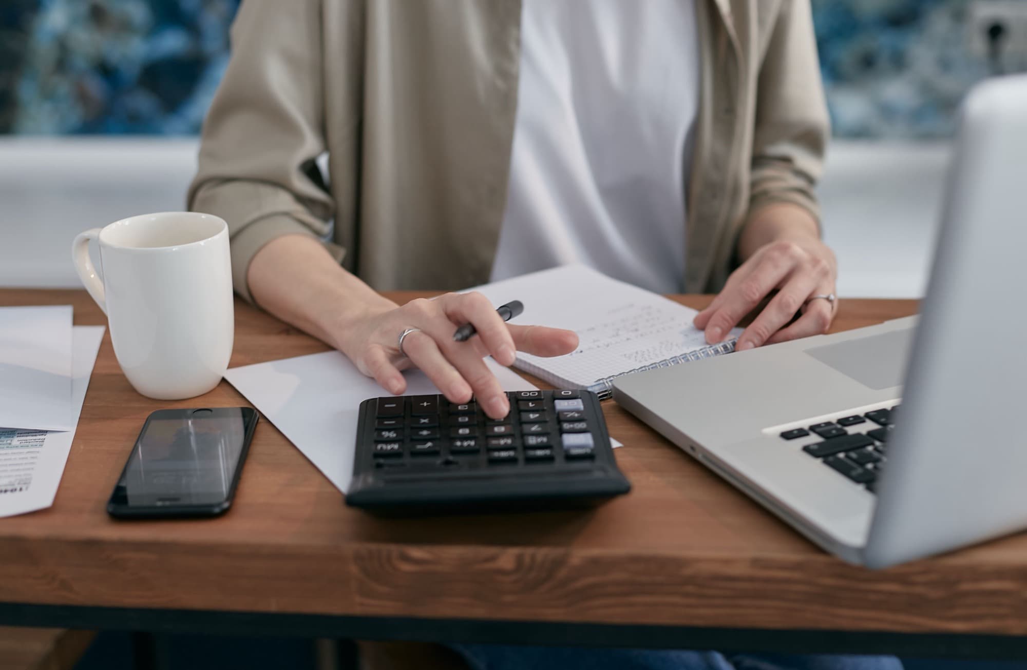A person at a wooden desk working a calculator next to a notebook, a laptop and a coffee mug