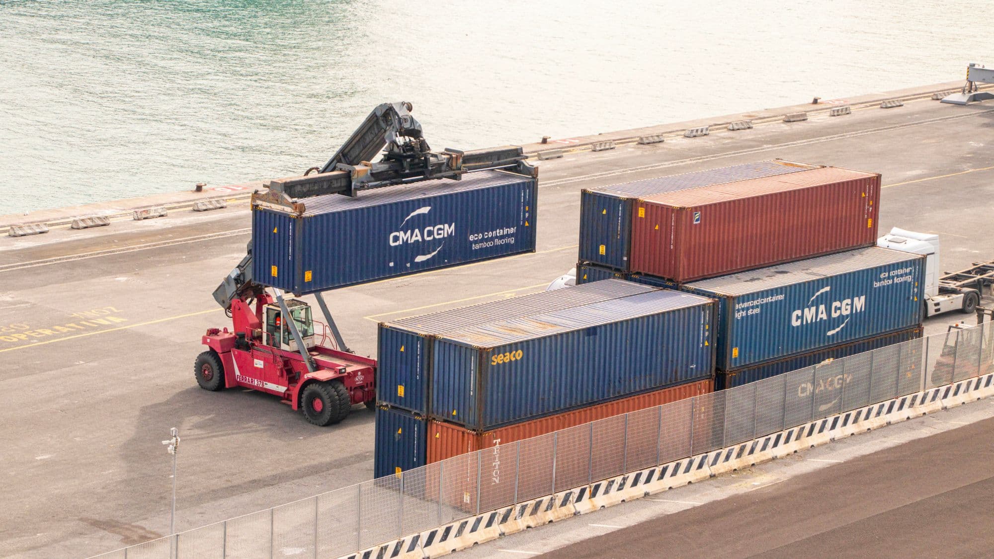 A reach stacker lifting a shipping container beside stacked containers on a quayside